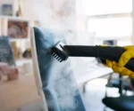 Sessel wird mittels eines Dampfreinigers von einer Person mit einem gelben Handschuh gereinigt. Im Hintergrund sind Umrisse eines Zimmers zu sehen.
